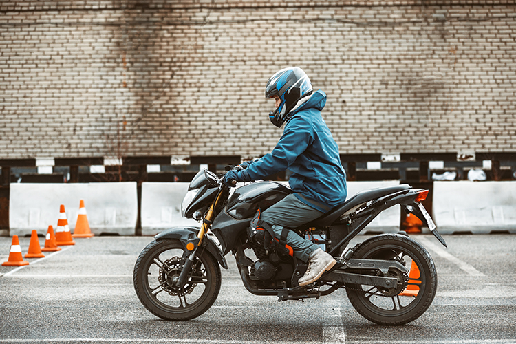 A young motorcyclist in a helmet on a black bike, riding round a car park