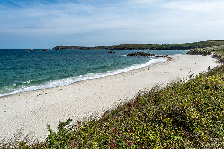 A photo of an empty white sand beach with blue water, Surrounded by grass. Isles of Scilly