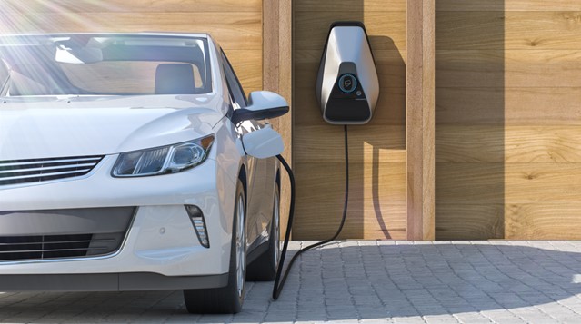 A white car being charged outside a house with a wooden fence and a built in charger on the wall