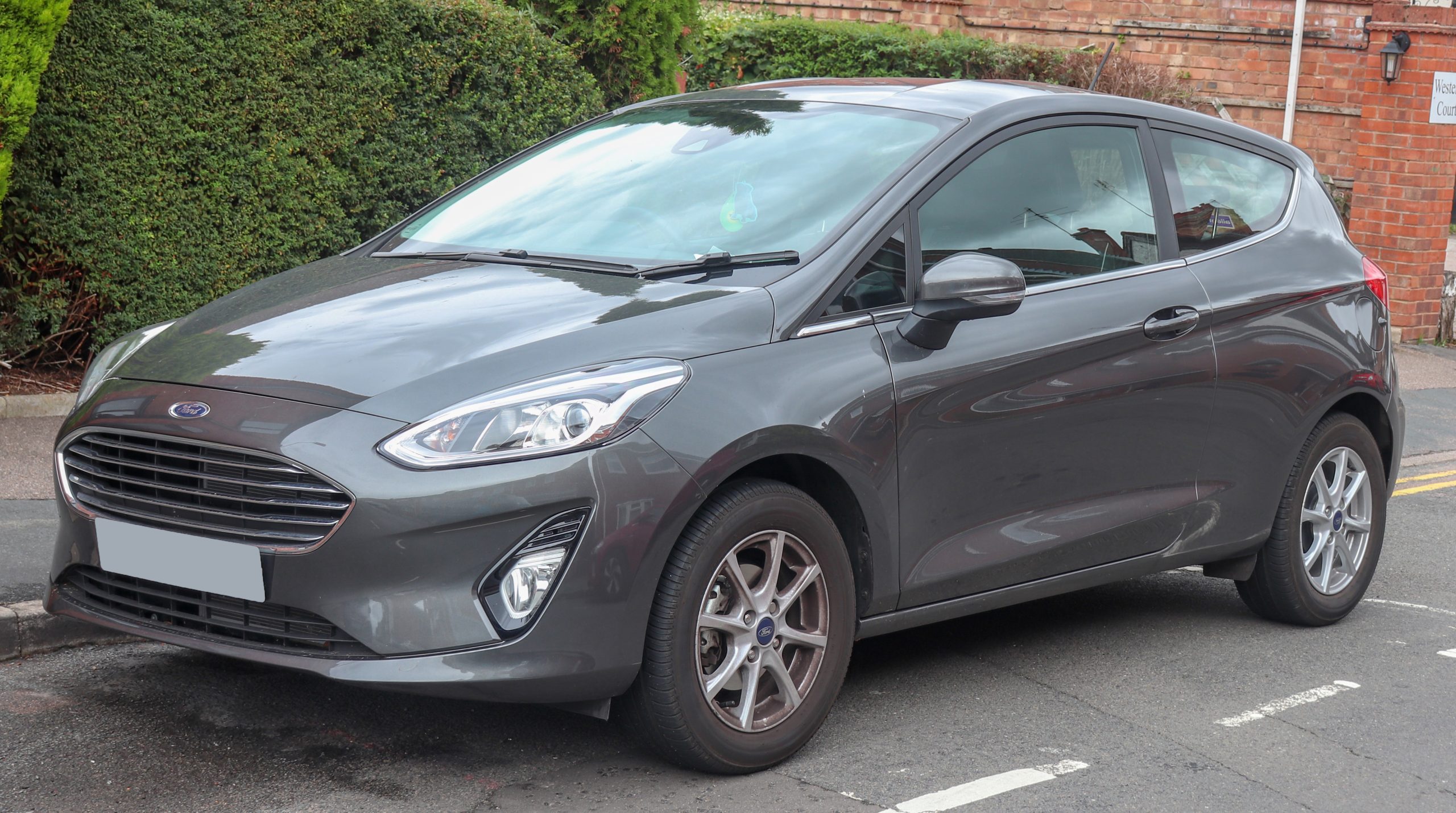 Grey ford fiesta zetec parked on side of road next to bushes and a brick wall