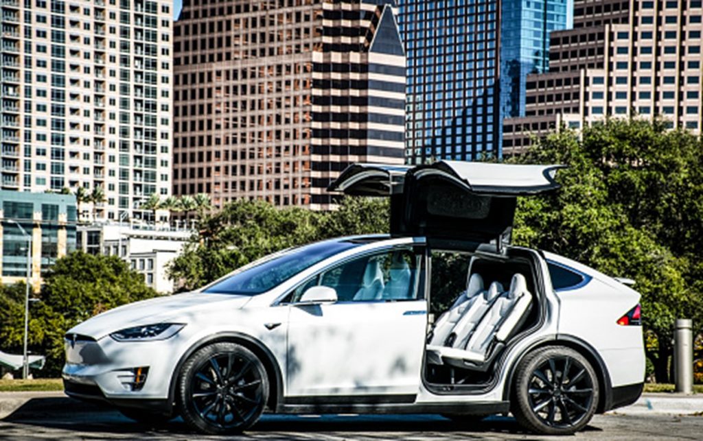 A white Tesla X with back wing doors open parked in front of high-rise buildings and greenery