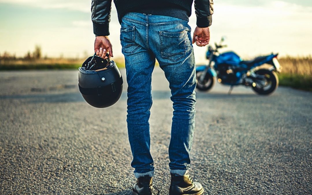 A low shot figure in blue jeans and a black leather jacket holding onto a biker helmet, walking towards a bike on a summers day