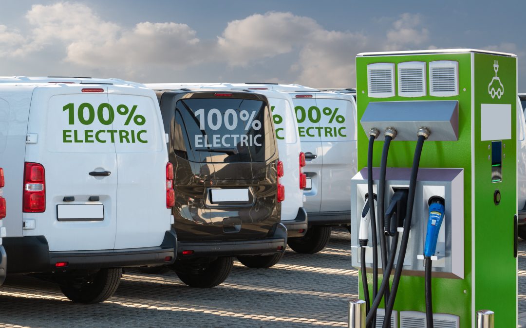 A white, a black and a white van rear facing with "100% Electric" parked next to a green EV charging point