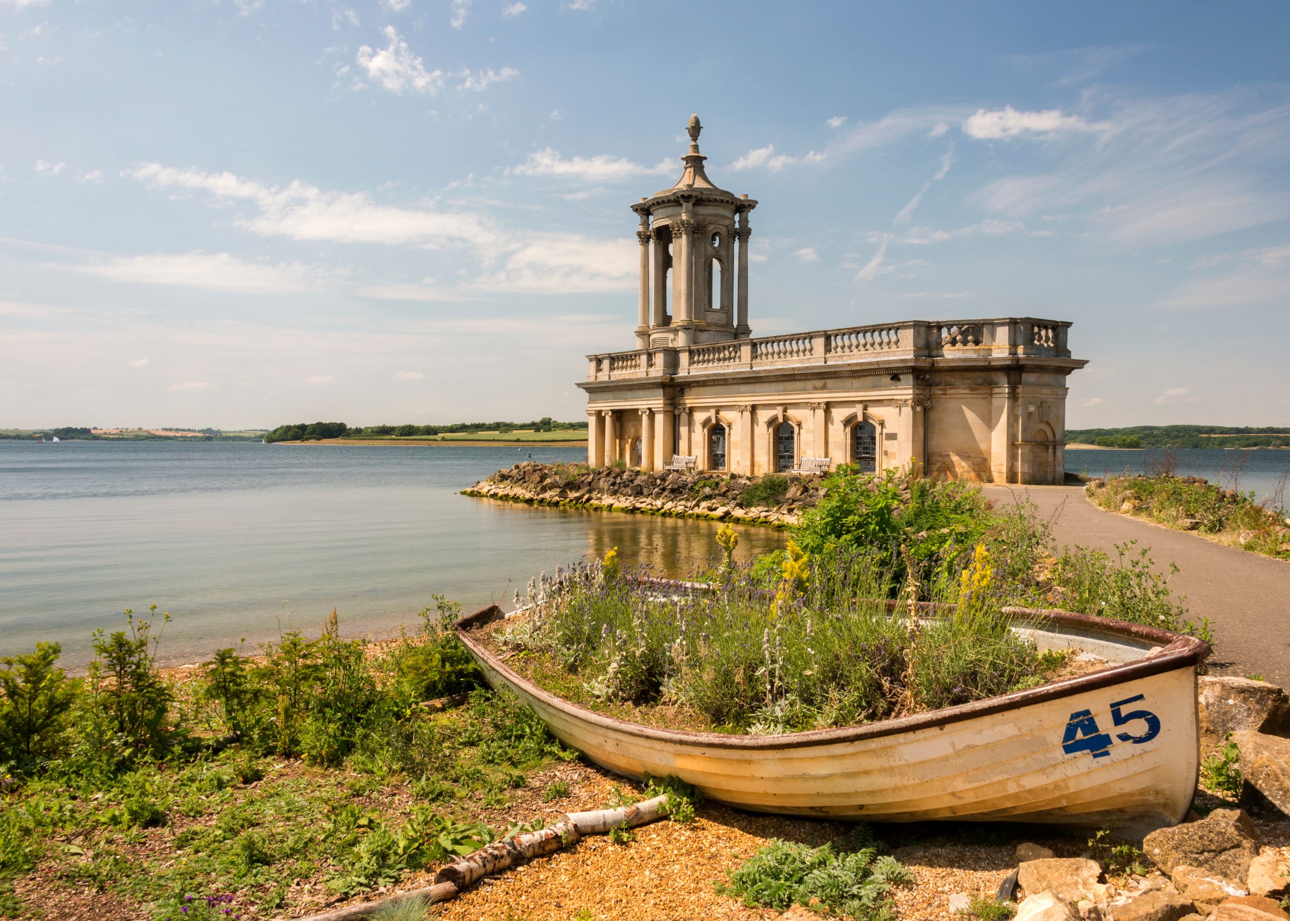 normanton church on rutland water. An old rowing boat filled with greenary on the banks of water. Behind is a small Church.