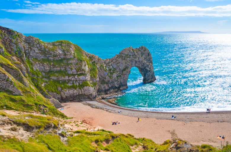 Sunny day at Durdle door. View from the top of a grassy cliff looking down on the beach and blue sea. Featuring Durdle door's famous limestone arch