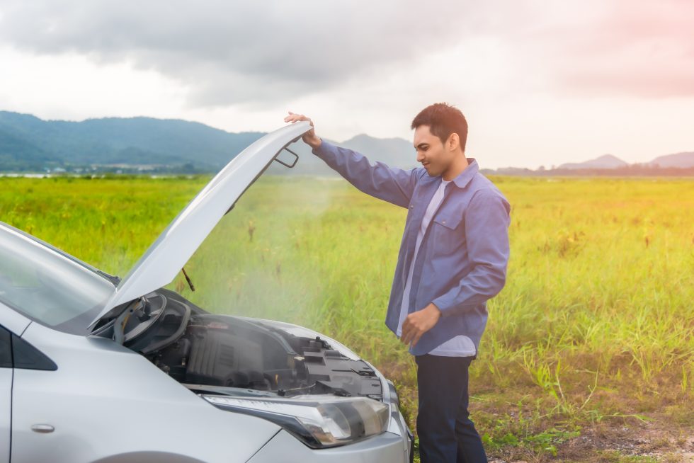 Male holding bonnet of silver car with smoke coming out of it in front of greenery