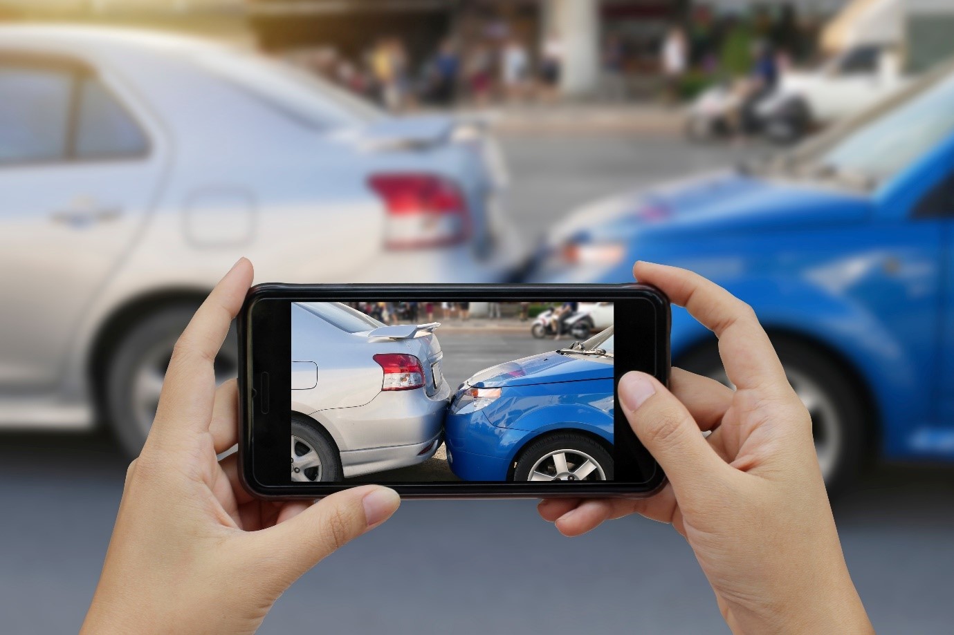 A Point of view shot looking at a landscape phone taking a picture of a car crash. A blue car crashed into a silver car