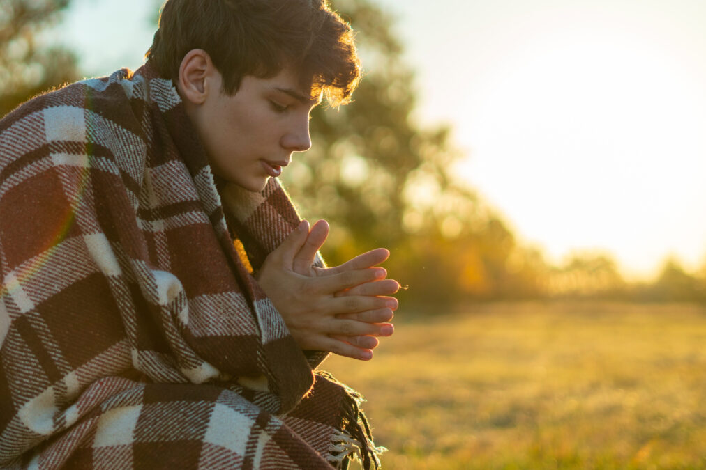 young frozen teenager covered by a warm blanket sitting on the grass field against the sun