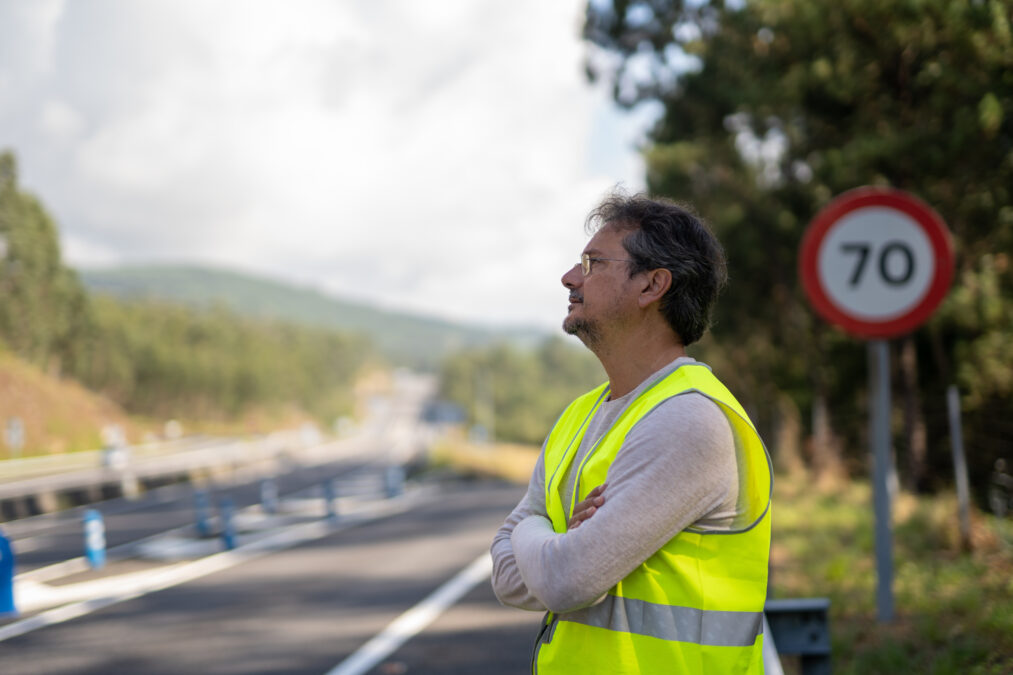 Caucasian man waiting a mechanic to come fix his broken down vehicle on the highway wearing a reflective vest.