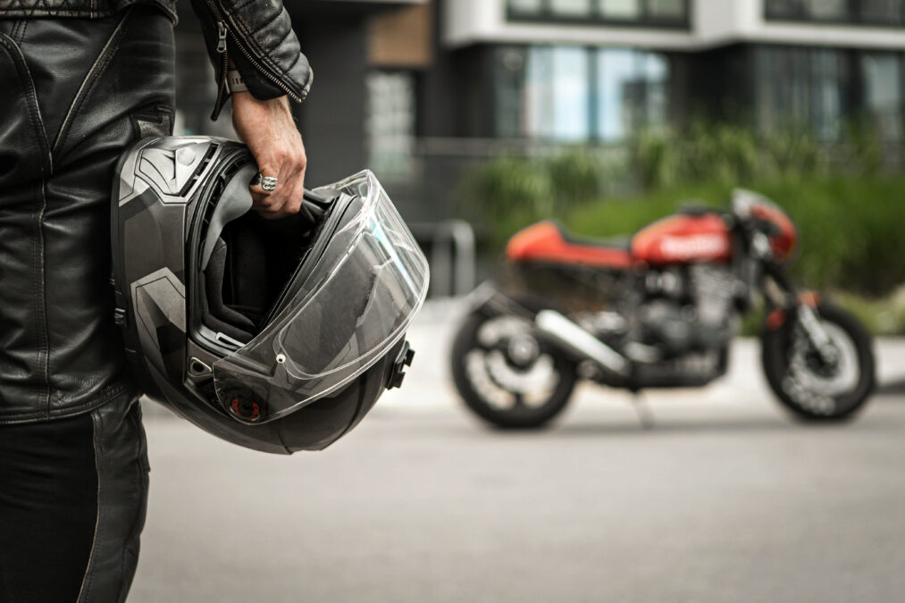 Male in all leathers holding a motorcycle helmet stood facing a red motorbike parked in front of a house.