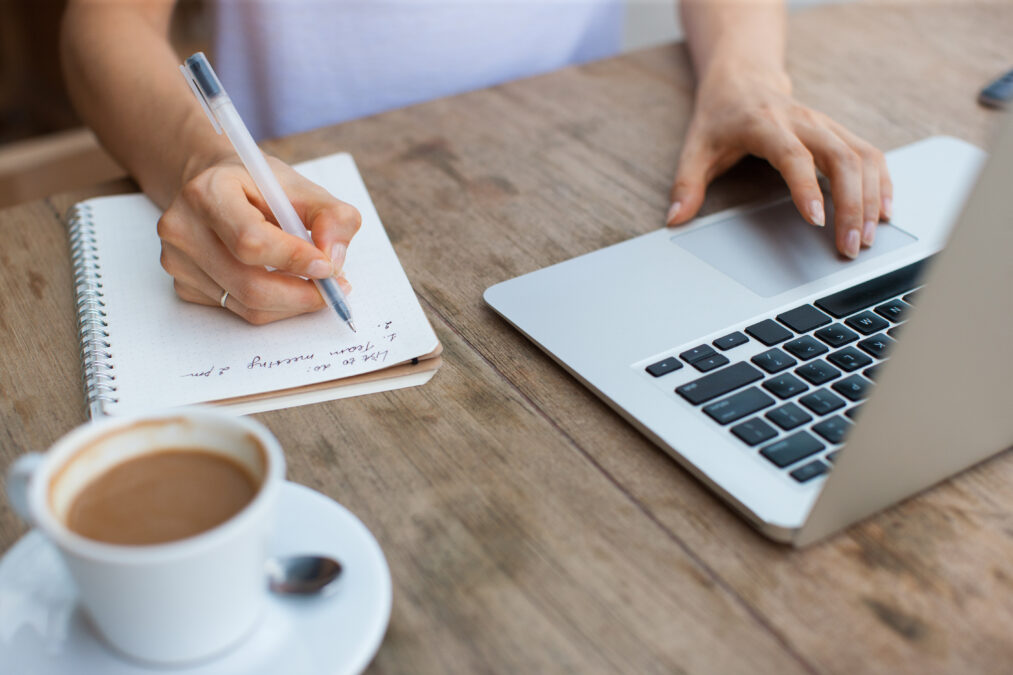 Cropped view of business woman hands working, typing on laptop computer and making notes in notebook at cafe table