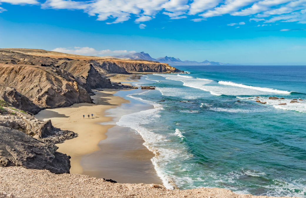 A sandy beach surrounded by costal rocks and 3 surfers walking down to the water with high waves. 