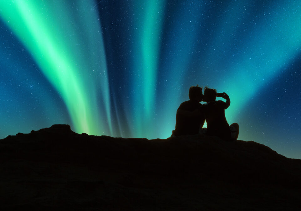 Couple kiss on a rock below the Green Aurora borealis shimmers over Reykjavik, Iceland.