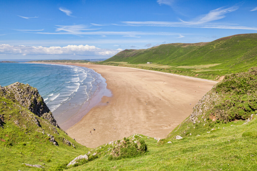 Greenery in the foreground and green hills in the background. Looking down on a sandy beach with blue waves 