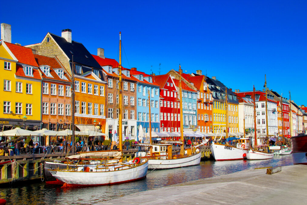 A row of colourful buildings alongside a  river with white sailing boats 