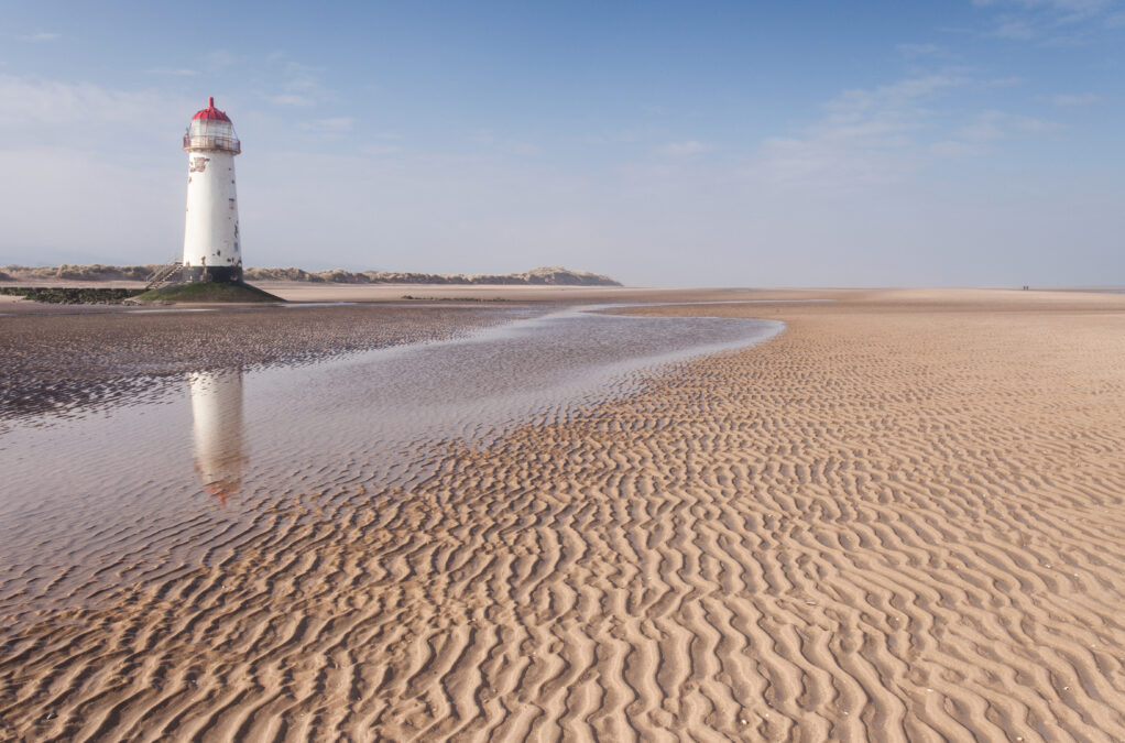 A rippled sandy beach with blue sky. To the left is q puddle in the sand and a white lighthouse with a red roof