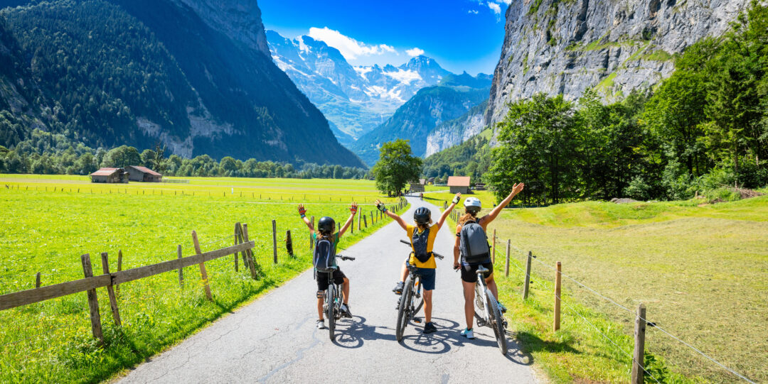 3 people including a child sat on their bikes with their arms in the air. on a cycle path surrounded by grass and the alps in the background 