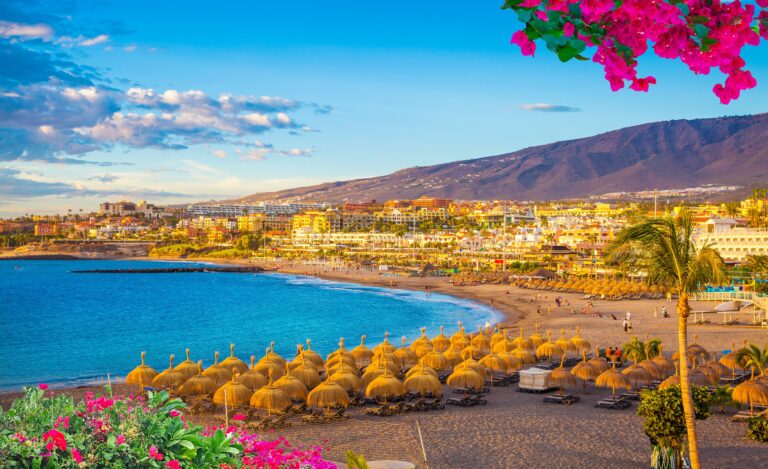 Coastal town at sunset with a sandy beach, straw parasols, and white buildings on a hillside.