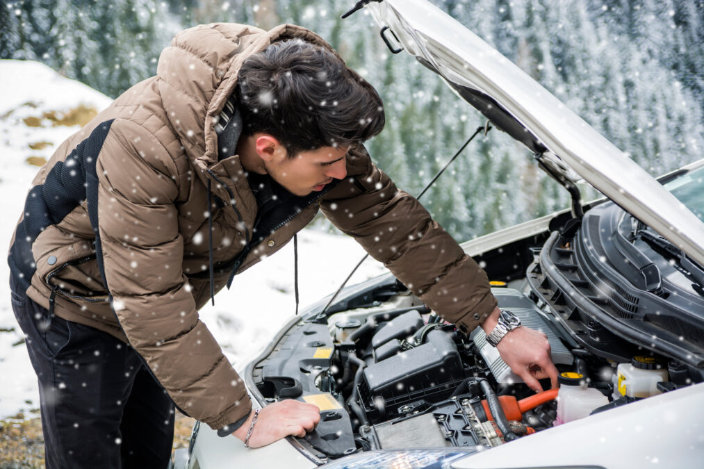 Young man near car with open hood inspecting engine in winter. Snowy forest on background