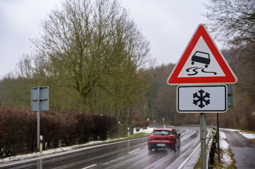 Car passing on a wet, mountain road in winter with a slippery road and ice warning sign showing a snowflake visible on the side.