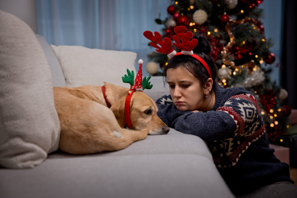 Sad lonely young woman sitting frustrated next to her yellow dog by couch at home during festive season, wearing Christmas antlers