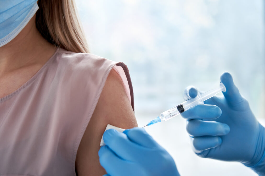 Doctor holding syringe making vaccination injection dose in shoulder of female patient wearing mask.